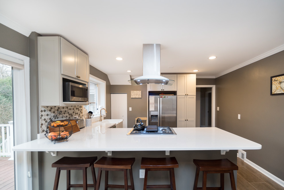 Mid-sized transitional l-shaped open plan kitchen in DC Metro with a farmhouse sink, shaker cabinets, white cabinets, quartz benchtops, grey splashback, porcelain splashback, stainless steel appliances, laminate floors and a peninsula.
