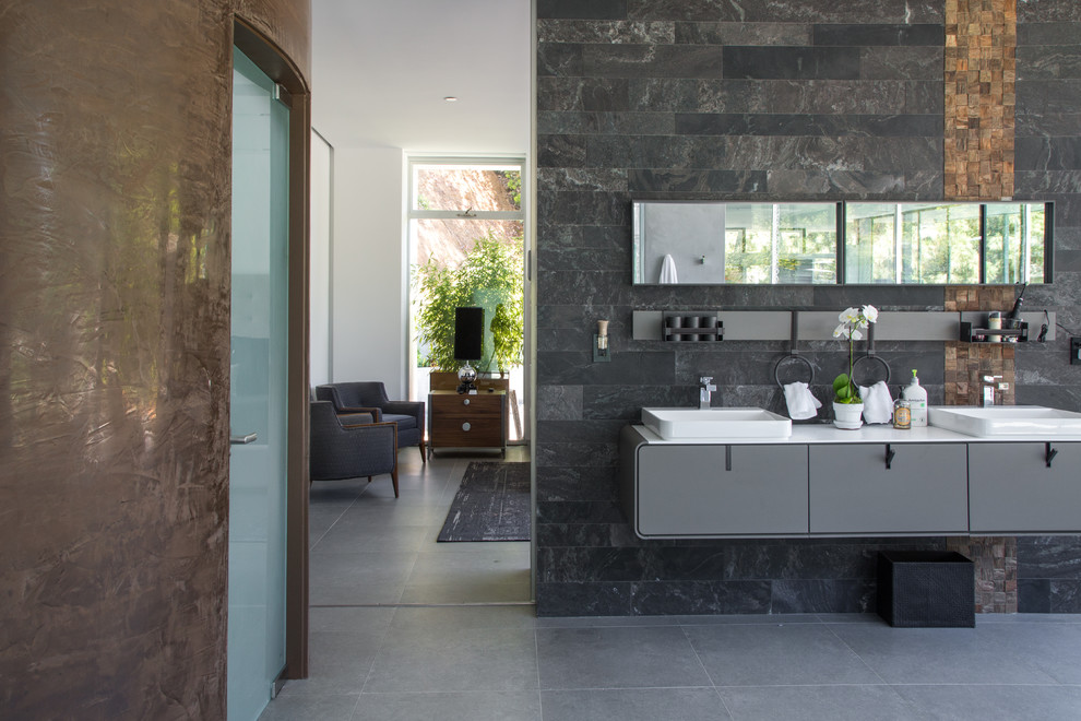 This is an example of a mid-sized modern master bathroom in Los Angeles with flat-panel cabinets, grey cabinets, a freestanding tub, an alcove shower, a two-piece toilet, black tile, gray tile, stone tile, white walls, concrete floors, an integrated sink, solid surface benchtops, grey floor and an open shower.