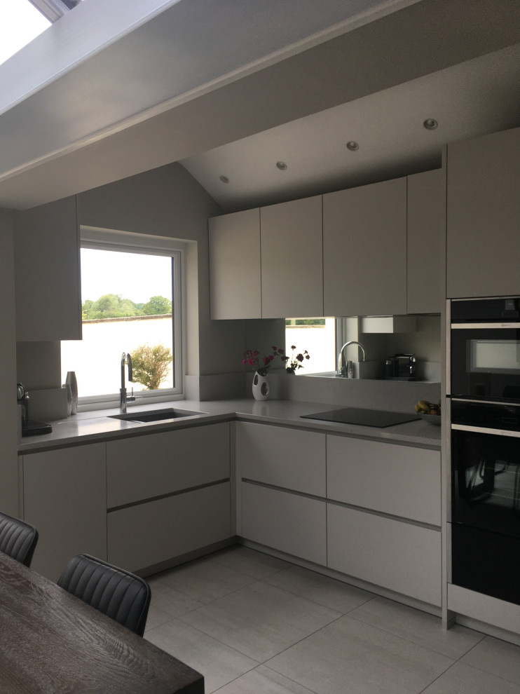 Small contemporary l-shaped eat-in kitchen in Manchester with an undermount sink, flat-panel cabinets, white cabinets, quartzite benchtops, metallic splashback, mirror splashback, black appliances, porcelain floors, no island, grey floor, white benchtop and vaulted.