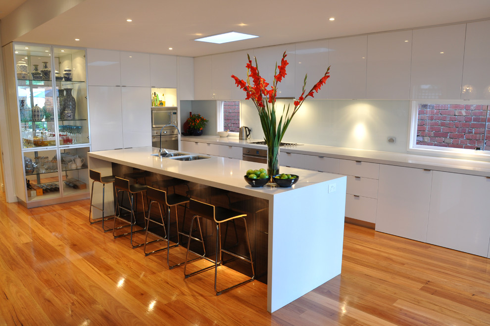 This is an example of a mid-sized modern l-shaped open plan kitchen in Geelong with an undermount sink, flat-panel cabinets, white cabinets, quartz benchtops, white splashback, glass sheet splashback, stainless steel appliances, light hardwood floors and with island.