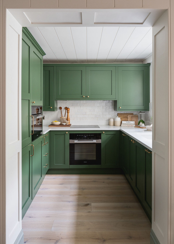 Design ideas for a small country eat-in kitchen in Sussex with recessed-panel cabinets, green cabinets, white splashback, ceramic splashback, panelled appliances, light hardwood floors, no island and white benchtop.