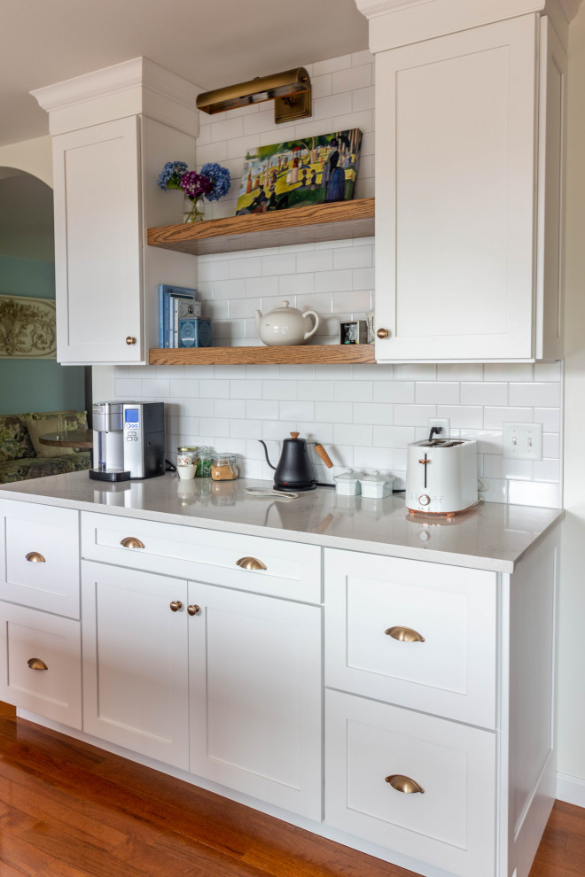 Example of a mid-sized transitional l-shaped medium tone wood floor and brown floor open concept kitchen design with an undermount sink, shaker cabinets, white cabinets, quartzite countertops, white backsplash, ceramic backsplash, white appliances, an island and white countertops