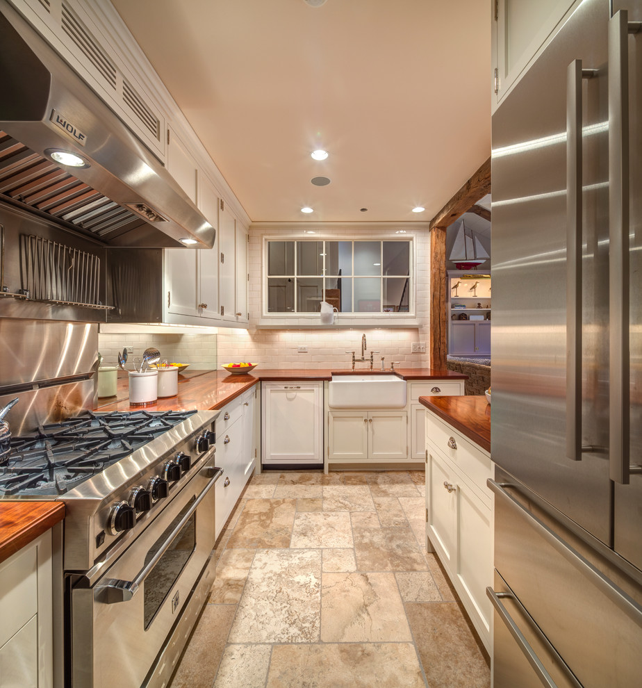 Mid-sized country l-shaped kitchen in Bridgeport with a farmhouse sink, recessed-panel cabinets, white cabinets, wood benchtops, white splashback, subway tile splashback, stainless steel appliances, a peninsula, limestone floors and beige floor.