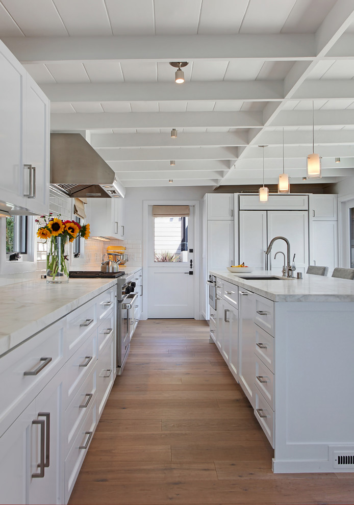 Inspiration for a beach style l-shaped kitchen in Orange County with an undermount sink, shaker cabinets, white cabinets, white splashback, panelled appliances, medium hardwood floors and with island.