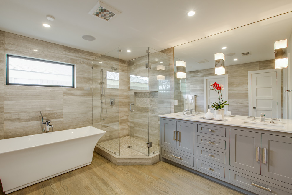 Photo of a mid-sized transitional master bathroom in Dallas with shaker cabinets, grey cabinets, a freestanding tub, a corner shower, gray tile, stone tile, grey walls, light hardwood floors, an undermount sink, engineered quartz benchtops and a hinged shower door.