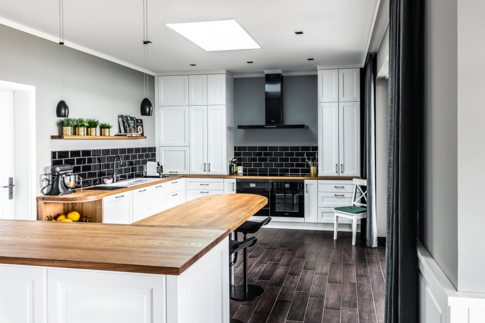 This is an example of a mid-sized industrial l-shaped open plan kitchen in Berlin with a double-bowl sink, beaded inset cabinets, white cabinets, wood benchtops, black splashback, ceramic splashback, black appliances, porcelain floors, a peninsula, brown floor and brown benchtop.