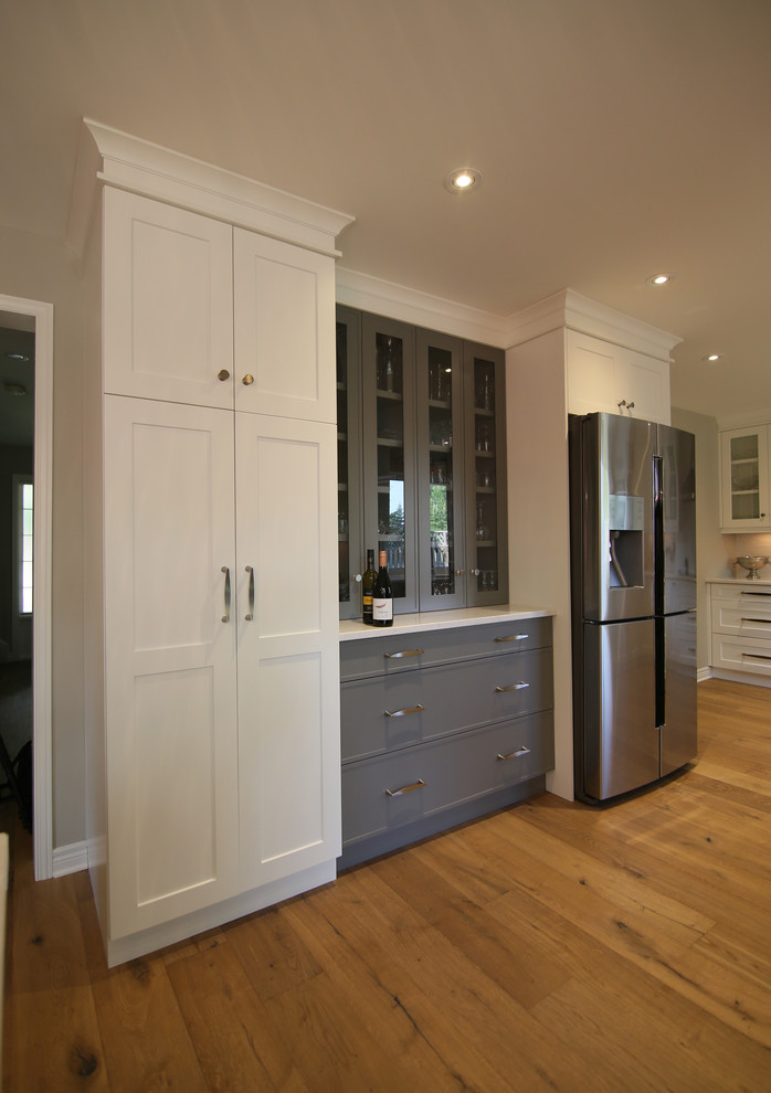 Example of a large transitional l-shaped medium tone wood floor and brown floor eat-in kitchen design with an undermount sink, shaker cabinets, white cabinets, stainless steel appliances, an island, solid surface countertops and white backsplash