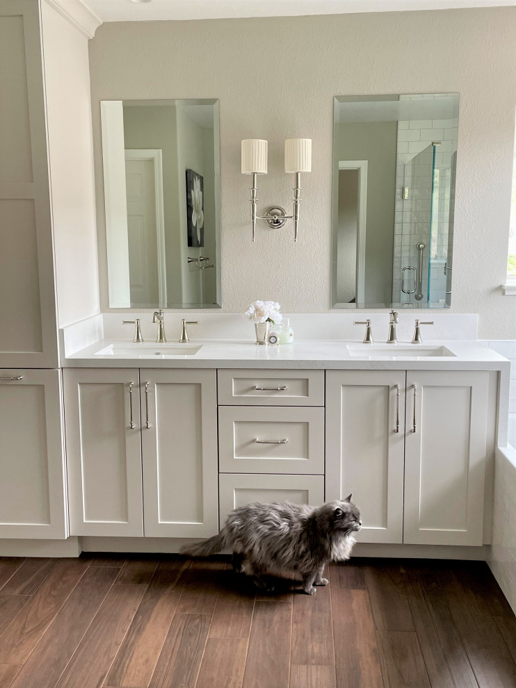 This is an example of a mid-sized transitional master bathroom in San Francisco with shaker cabinets, white cabinets, an undermount tub, a corner shower, white tile, ceramic tile, white walls, laminate floors, an undermount sink, engineered quartz benchtops, brown floor, a hinged shower door, white benchtops, a shower seat, a double vanity and a built-in vanity.