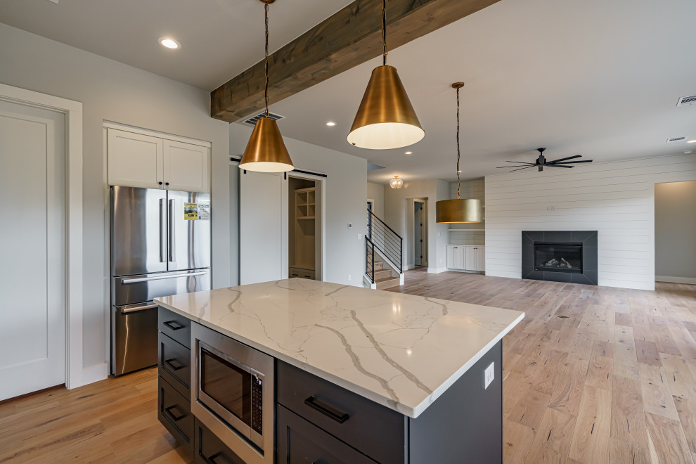 This is an example of a country l-shaped open plan kitchen in Austin with an undermount sink, shaker cabinets, white cabinets, marble benchtops, white splashback, stainless steel appliances, light hardwood floors, with island, white benchtop and exposed beam.