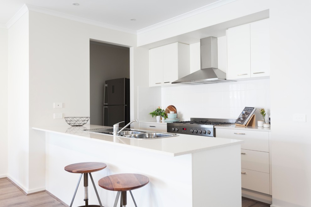 This is an example of a contemporary galley kitchen in Melbourne with a drop-in sink, flat-panel cabinets, white cabinets, white splashback, stainless steel appliances, medium hardwood floors, a peninsula, brown floor and white benchtop.