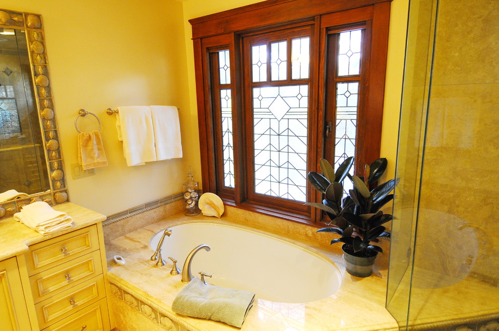 Photo of a mid-sized tropical master bathroom in San Diego with recessed-panel cabinets, beige cabinets, an undermount tub, a corner shower, a two-piece toilet, yellow tile, travertine, beige walls, light hardwood floors, an undermount sink, limestone benchtops, brown floor and a hinged shower door.