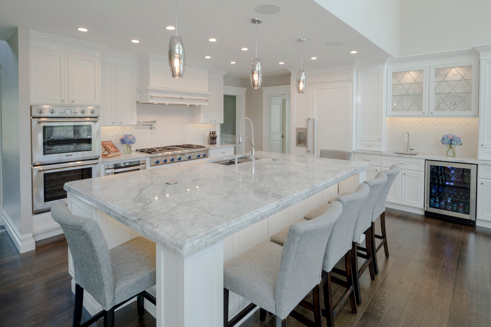 Large modern l-shaped open plan kitchen in Boston with an undermount sink, beaded inset cabinets, white cabinets, marble benchtops, white splashback, ceramic splashback, stainless steel appliances, dark hardwood floors, with island and brown floor.
