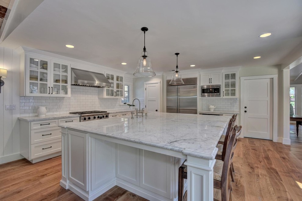Photo of a mid-sized country l-shaped open plan kitchen in San Francisco with a farmhouse sink, recessed-panel cabinets, white cabinets, marble benchtops, white splashback, subway tile splashback, stainless steel appliances, medium hardwood floors and with island.