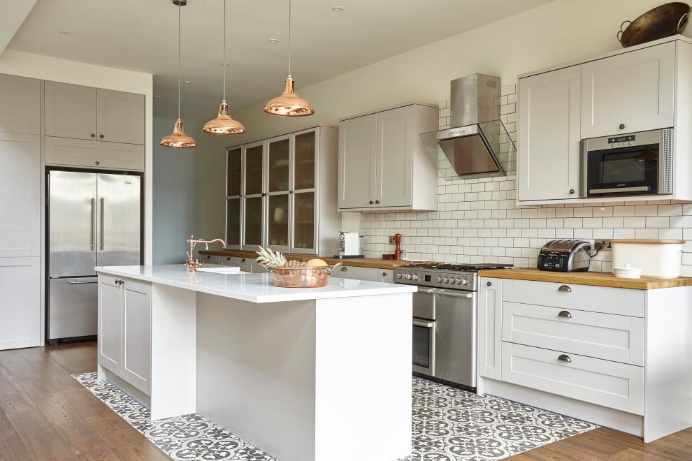 Transitional galley kitchen in London with a farmhouse sink, shaker cabinets, white cabinets, wood benchtops, white splashback, subway tile splashback, stainless steel appliances, with island, multi-coloured floor and brown benchtop.
