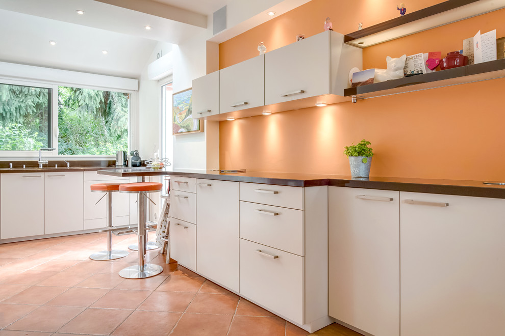Design ideas for a contemporary kitchen in Paris with flat-panel cabinets, orange splashback, terra-cotta floors, a peninsula, orange floor, brown benchtop, stainless steel appliances, a double-bowl sink, beige cabinets and wood benchtops.