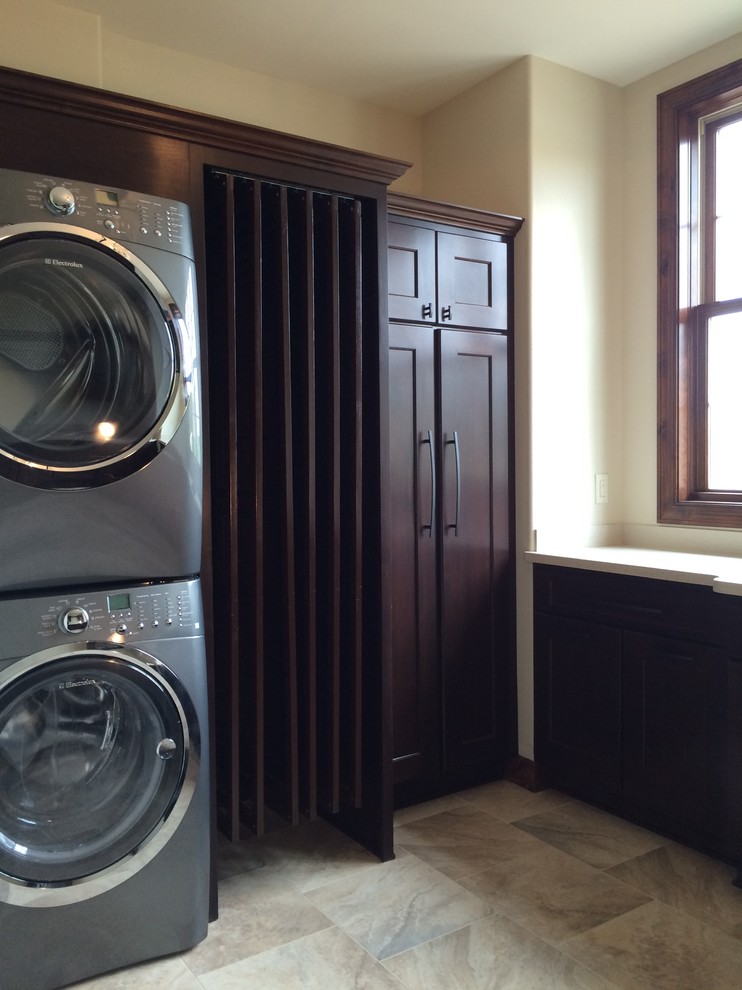 Arts and crafts laundry room in Milwaukee with dark wood cabinets, white walls, ceramic floors and a stacked washer and dryer.
