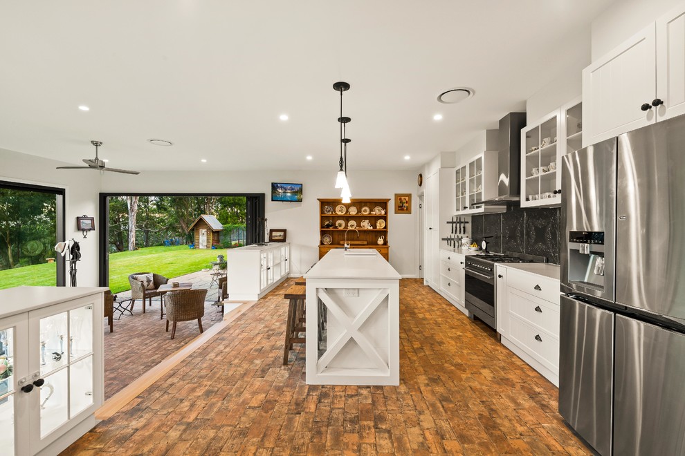Photo of a country galley open plan kitchen in Other with shaker cabinets, white cabinets, black splashback, stainless steel appliances, brick floors, with island, brown floor and white benchtop.