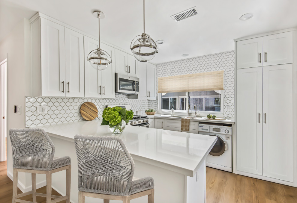 Design ideas for a transitional u-shaped kitchen in Los Angeles with an undermount sink, shaker cabinets, white cabinets, white splashback, white appliances, medium hardwood floors, a peninsula, brown floor and white benchtop.