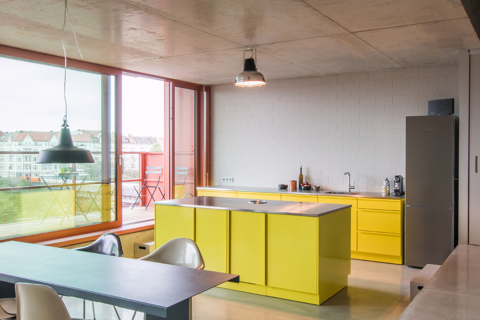 This is an example of a contemporary single-wall eat-in kitchen in Berlin with a single-bowl sink, flat-panel cabinets, yellow cabinets, stainless steel benchtops, white splashback, stainless steel appliances, concrete floors and with island.
