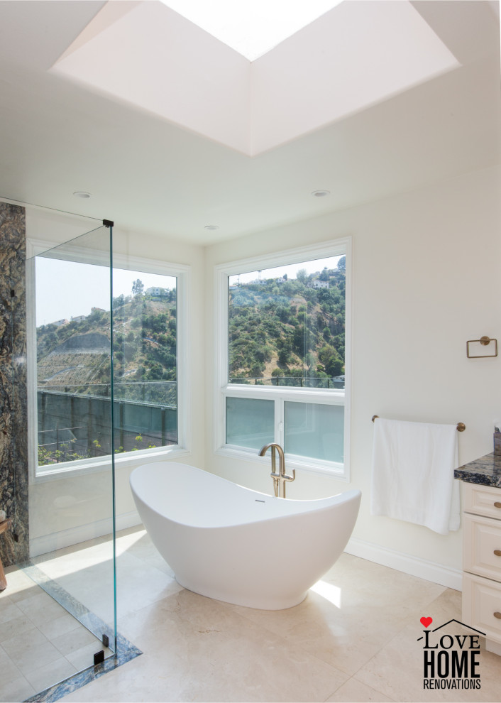 This is an example of a large mediterranean master bathroom in Los Angeles with beaded inset cabinets, beige cabinets, a freestanding tub, a curbless shower, a one-piece toilet, multi-coloured tile, stone slab, white walls, travertine floors, a drop-in sink, granite benchtops, beige floor, a hinged shower door, multi-coloured benchtops, an enclosed toilet, a double vanity and a built-in vanity.