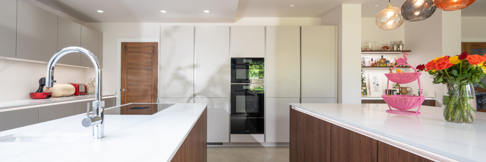 Photo of a large industrial galley open plan kitchen in Hertfordshire with flat-panel cabinets, brown cabinets, quartzite benchtops, black appliances, with island and white benchtop.