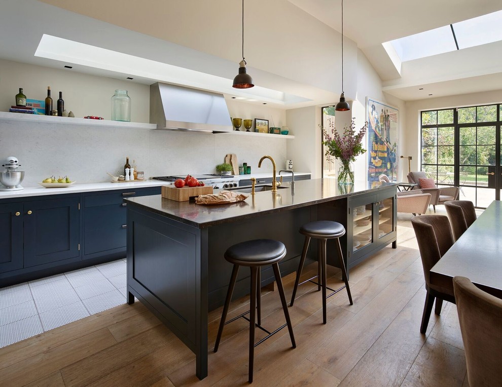 This is an example of a mid-sized contemporary single-wall open plan kitchen in Other with recessed-panel cabinets, blue cabinets, marble benchtops, white splashback, marble splashback, stainless steel appliances, light hardwood floors, with island and brown floor.