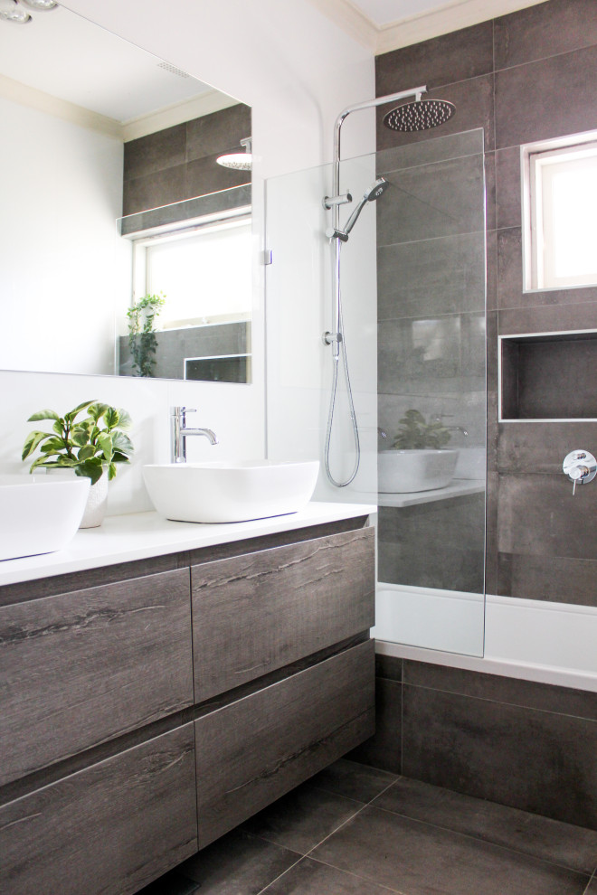 This is an example of a mid-sized contemporary kids bathroom in Perth with flat-panel cabinets, dark wood cabinets, a drop-in tub, a shower/bathtub combo, a one-piece toilet, white tile, ceramic tile, white walls, porcelain floors, a vessel sink, engineered quartz benchtops, grey floor, a hinged shower door, white benchtops, a niche and a floating vanity.