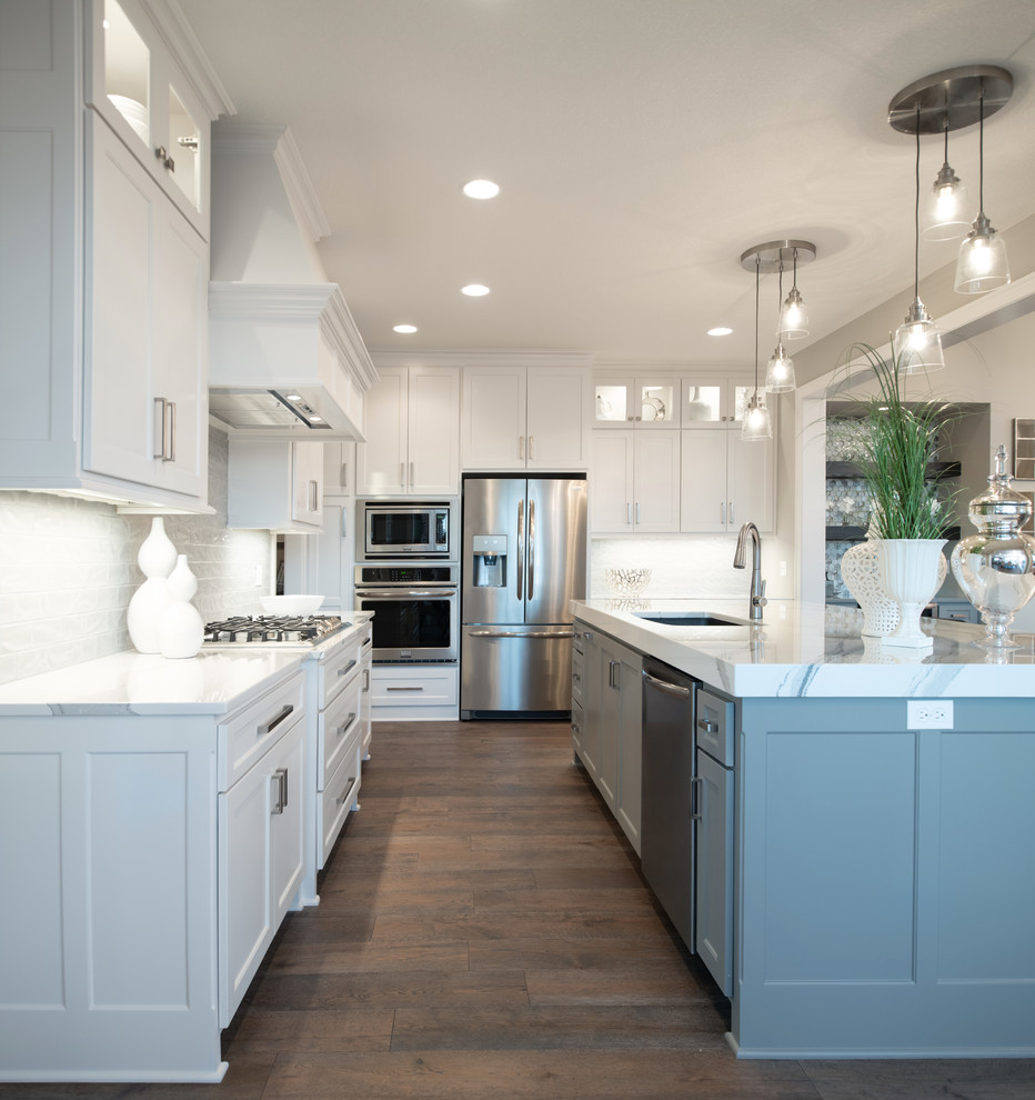 Photo of an arts and crafts l-shaped open plan kitchen in Kansas City with a drop-in sink, white cabinets, white splashback, stainless steel appliances, medium hardwood floors, with island, brown floor and white benchtop.