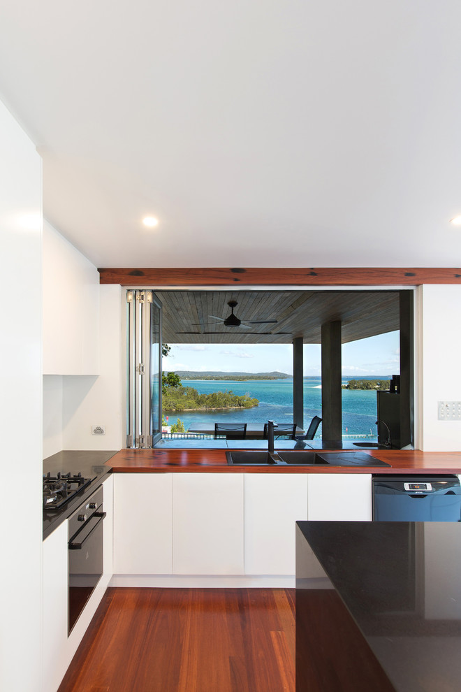 Design ideas for a mid-sized contemporary l-shaped open plan kitchen in Newcastle - Maitland with a drop-in sink, flat-panel cabinets, white cabinets, wood benchtops, white splashback, glass sheet splashback, black appliances, medium hardwood floors and with island.