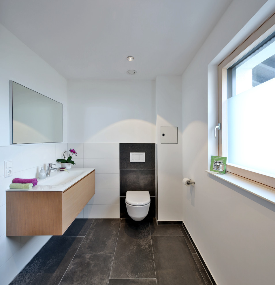 Photo of a mid-sized contemporary powder room in Munich with flat-panel cabinets, light wood cabinets, a two-piece toilet, black tile, white tile, cement tile, white walls, cement tiles, an integrated sink and black floor.