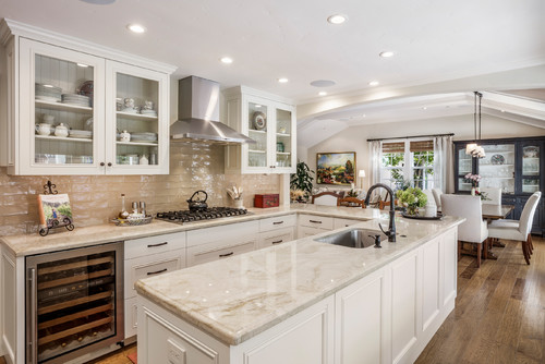 U-shape kitchen with white Shaker cabinets, marble-look countertops, glass-front uppers, stainless hood, wine cooler, and a central island sink in an open-plan space