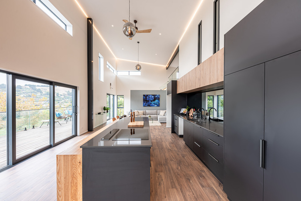 Inspiration for a small urban galley open plan kitchen in Christchurch with a single-bowl sink, black cabinets, composite countertops, mirror splashback, stainless steel appliances, medium hardwood flooring, an island, brown floors, grey worktops and a vaulted ceiling.