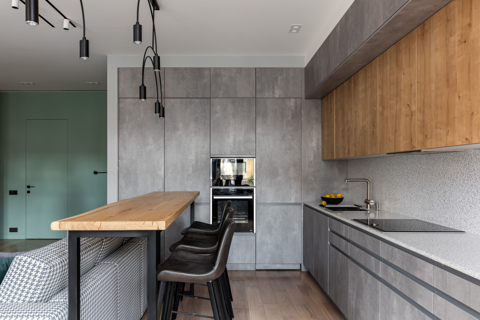 Mid-sized contemporary l-shaped eat-in kitchen in Saint Petersburg with a single-bowl sink, flat-panel cabinets, grey cabinets, solid surface benchtops, grey splashback, engineered quartz splashback, black appliances, medium hardwood floors, with island, brown floor and grey benchtop.