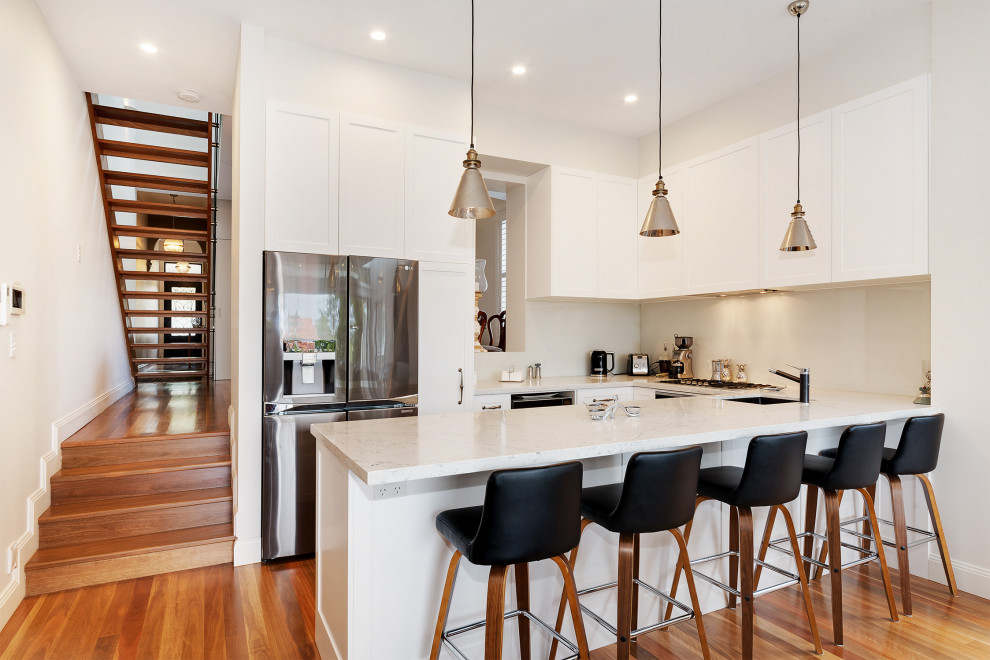 Inspiration for a mid-sized transitional u-shaped open plan kitchen in Sydney with a drop-in sink, shaker cabinets, white cabinets, quartz benchtops, white splashback, glass sheet splashback, stainless steel appliances, medium hardwood floors, with island, brown floor and white benchtop.