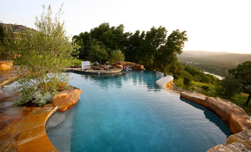 Large traditional backyard custom-shaped infinity pool in Austin with a water feature and natural stone pavers.