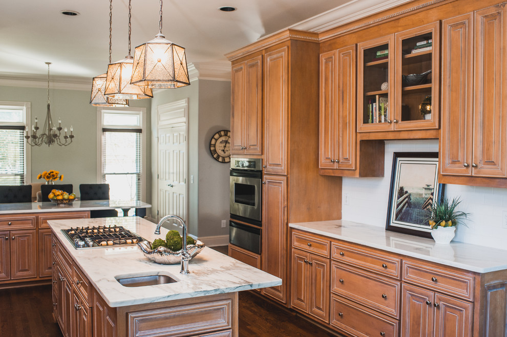 Mid-sized transitional u-shaped eat-in kitchen in Nashville with marble benchtops, white splashback, subway tile splashback, stainless steel appliances, with island, an undermount sink, raised-panel cabinets, brown cabinets, dark hardwood floors, brown floor and white benchtop.