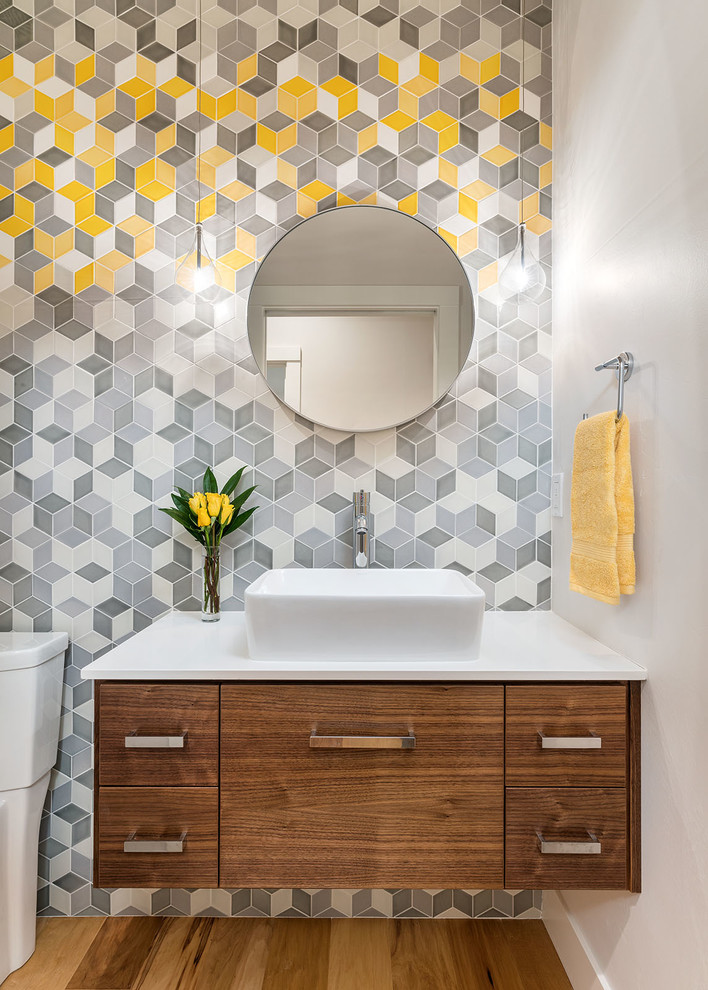 This is an example of a small contemporary powder room in Other with multi-coloured walls, a vessel sink, brown floor, flat-panel cabinets, medium wood cabinets, a two-piece toilet, gray tile, ceramic tile, light hardwood floors, engineered quartz benchtops and white benchtops.