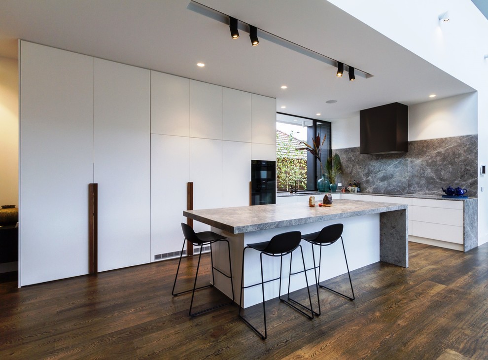 Large modern l-shaped open plan kitchen in Melbourne with an undermount sink, flat-panel cabinets, white cabinets, grey splashback, marble splashback, with island, brown floor, grey benchtop, panelled appliances, dark hardwood floors and limestone benchtops.