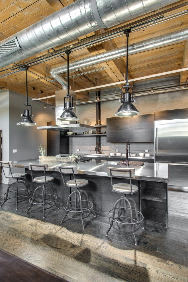 This is an example of a mid-sized industrial galley open plan kitchen in Chicago with a double-bowl sink, flat-panel cabinets, grey cabinets, stainless steel benchtops, grey splashback, stainless steel appliances, light hardwood floors and with island.