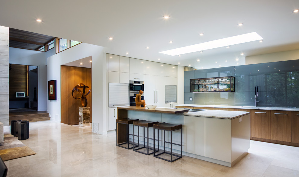 Photo of a large single-wall open plan kitchen in Calgary with a double-bowl sink, flat-panel cabinets, white cabinets, limestone benchtops, grey splashback, glass sheet splashback, stainless steel appliances, marble floors, with island and beige floor.