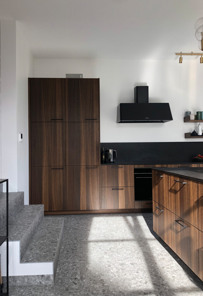 This is an example of an expansive midcentury galley separate kitchen in Munich with an undermount sink, flat-panel cabinets, dark wood cabinets, granite benchtops, black splashback, granite splashback, black appliances, terrazzo floors, with island, grey floor and black benchtop.