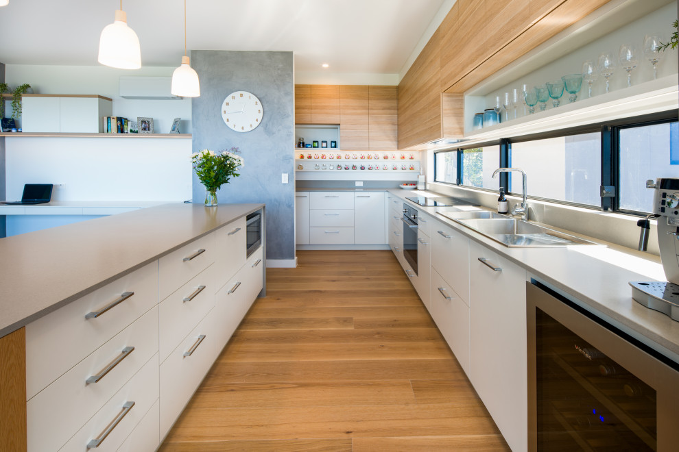 This is an example of an expansive contemporary u-shaped kitchen in Sunshine Coast with a double-bowl sink, flat-panel cabinets, white cabinets, quartz benchtops, grey splashback, stone slab splashback, stainless steel appliances, with island, brown floor, grey benchtop and medium hardwood floors.
