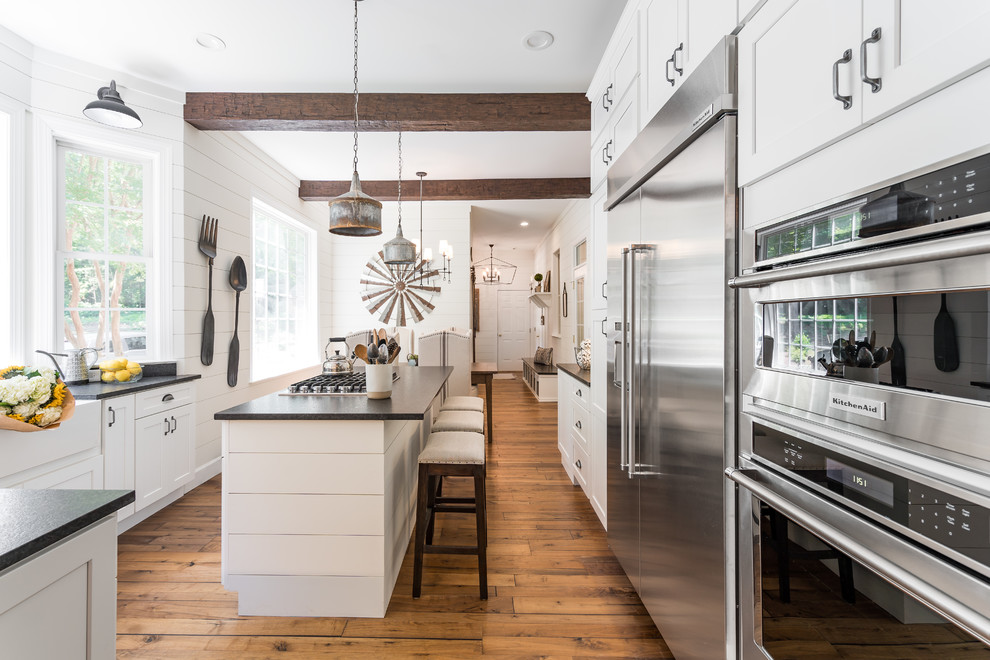 Country u-shaped eat-in kitchen in Raleigh with a farmhouse sink, shaker cabinets, white cabinets, stainless steel appliances, medium hardwood floors, with island, brown floor and black benchtop.