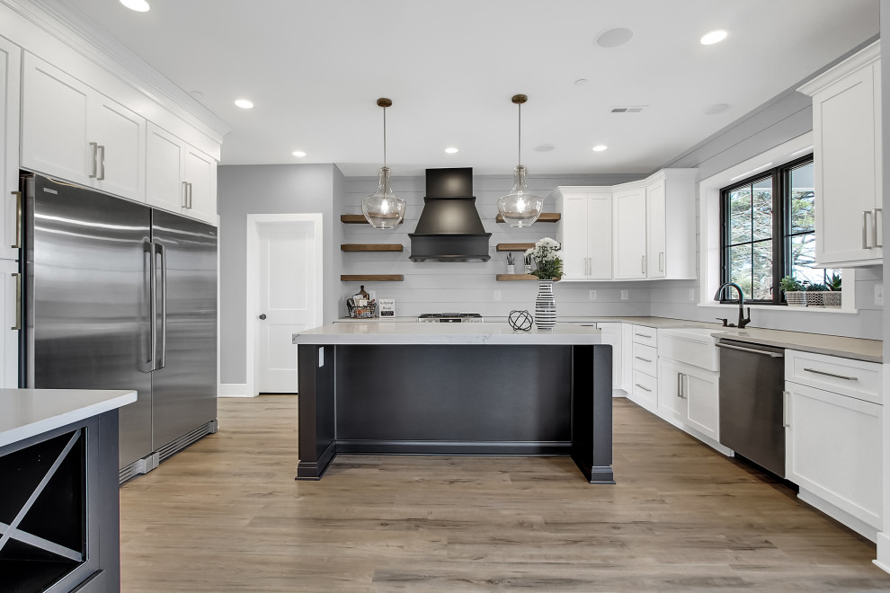 Large country l-shaped eat-in kitchen in Baltimore with with island, a farmhouse sink, grey splashback, shiplap splashback, stainless steel appliances, medium hardwood floors, shaker cabinets and white cabinets.