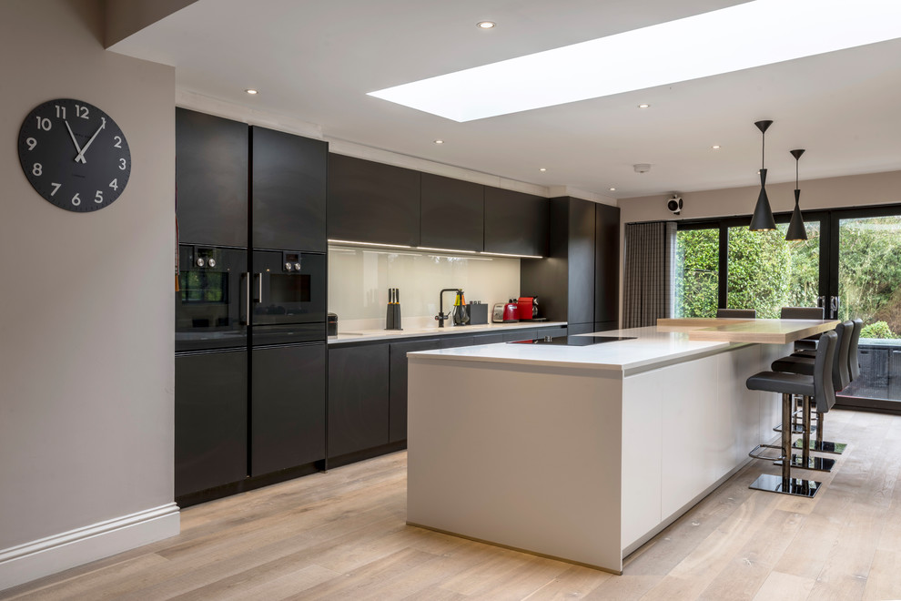 Large contemporary l-shaped open plan kitchen in Cheshire with a drop-in sink, flat-panel cabinets, solid surface benchtops, white splashback, glass sheet splashback, black appliances, light hardwood floors and with island.