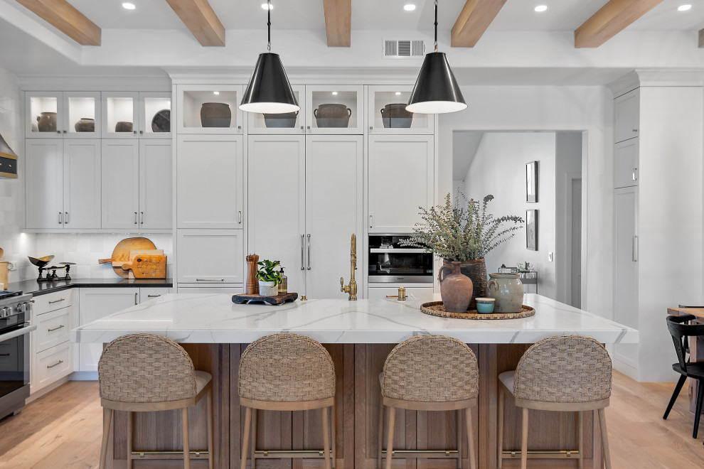 Medium sized classic l-shaped open plan kitchen in Los Angeles with a belfast sink, shaker cabinets, white cabinets, engineered stone countertops, white splashback, ceramic splashback, stainless steel appliances, light hardwood flooring, an island, white worktops and exposed beams.