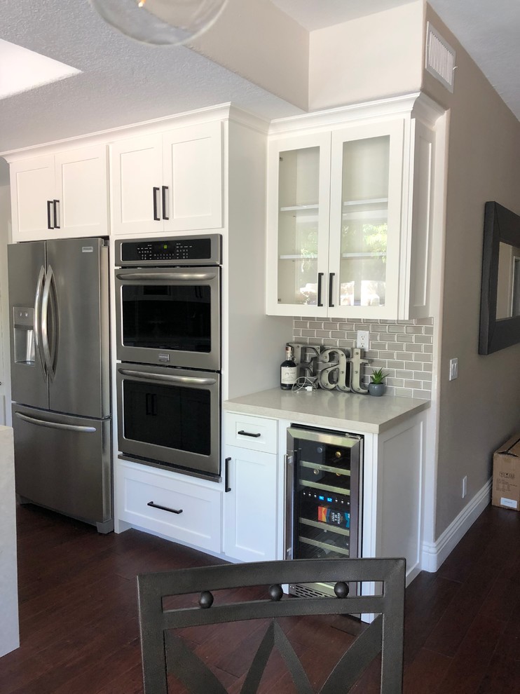Mid-sized country u-shaped eat-in kitchen in Orange County with a farmhouse sink, shaker cabinets, white cabinets, quartzite benchtops, grey splashback, subway tile splashback, stainless steel appliances, dark hardwood floors, with island, multi-coloured floor and beige benchtop.