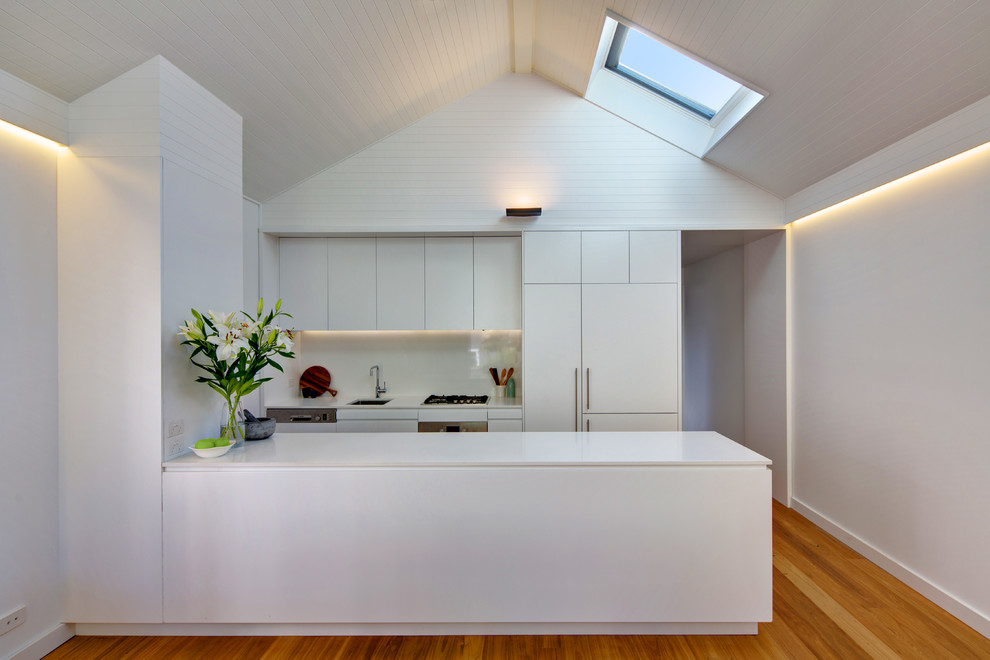This is an example of a mid-sized contemporary galley open plan kitchen in Sydney with an undermount sink, flat-panel cabinets, white cabinets, quartz benchtops, white splashback, glass sheet splashback, stainless steel appliances, medium hardwood floors and a peninsula.