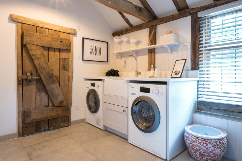 This is an example of a large country single-wall laundry room in Kent with a farmhouse sink, shaker cabinets, white cabinets, quartzite benchtops, white walls, grey floor, white benchtop, a side-by-side washer and dryer and exposed beam.