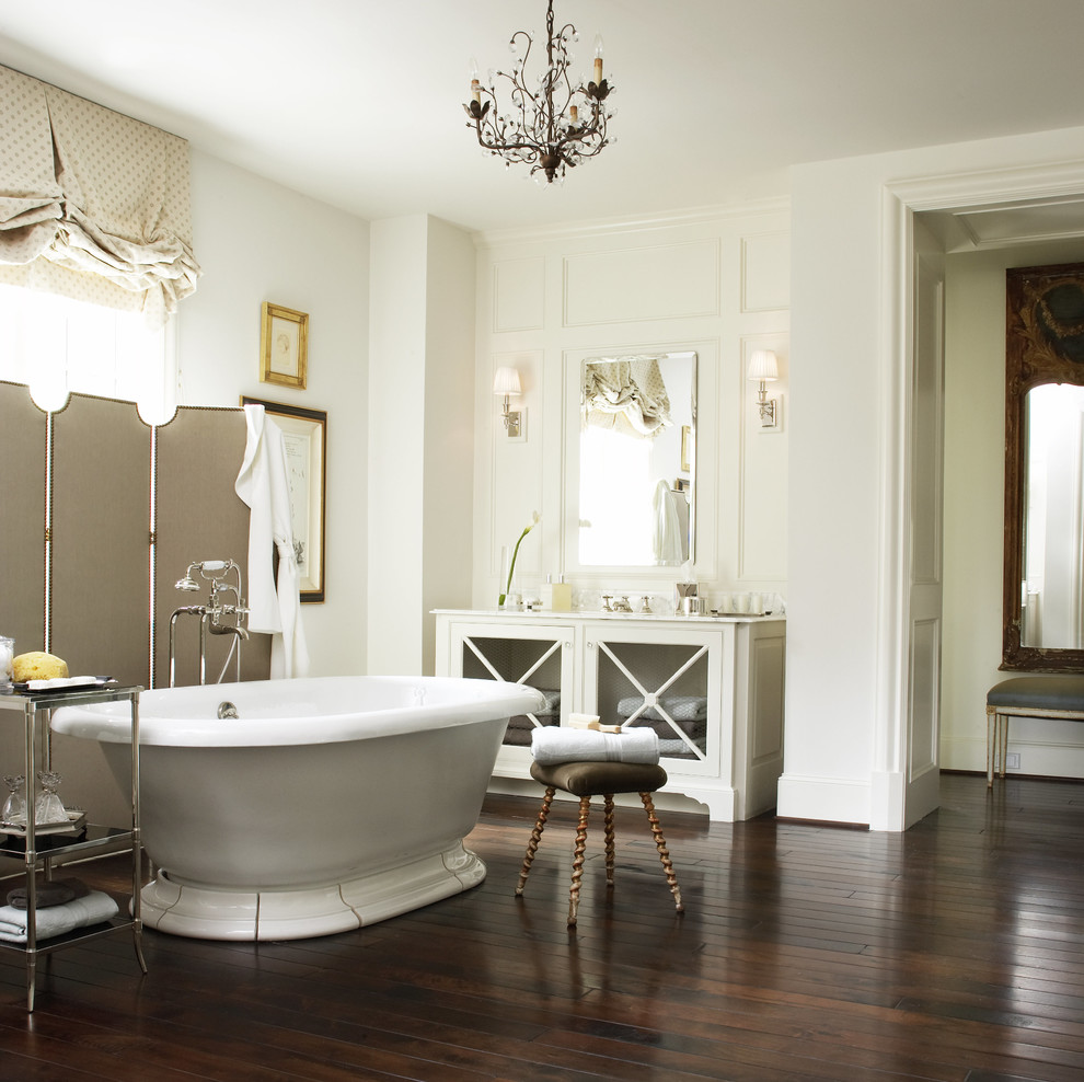This is an example of a mid-sized traditional master bathroom in Houston with a freestanding tub, glass-front cabinets, white cabinets, white walls, dark hardwood floors, an undermount sink, solid surface benchtops and brown floor.